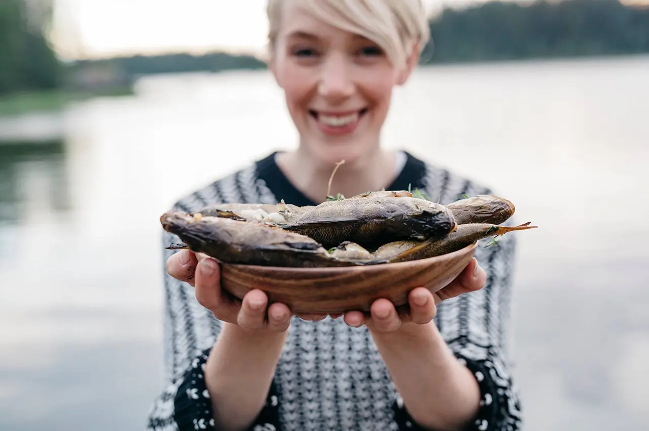 smiling woman offers a plate of fresh cooked fish. photo: Elina Manninen / Keksit